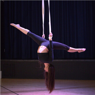 Personne suspendue à l’envers sur des sangles aériennes en coton Prodigy, tenant les sangles et les jambes étendues, vêtue d’un ensemble noir; scène de spectacle avec rideaux noirs en arrière-plan.