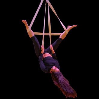 Femme en position d'acrobaties suspendue aux sangles aériennes en coton Prodigy, vêtu d'un ensemble noir, les bras tendus et les pieds écartés, sur fond noir.