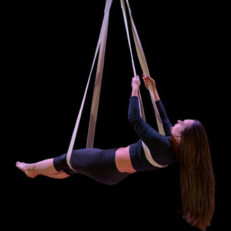 Femme en position suspendue sur des sangles aériennes en coton, portant un haut noir et un legging. Elle tient les sangles avec les mains, la tête légèrement incliné en arrière, les cheveux longs tombant autour d'elle. Fond noir.