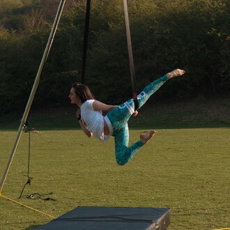 Jeune femme en train de s'exercer sur des sangles aériennes, suspendue dans les airs avec une jambe tendue. Elle porte un t-shirt blanc et un legging turquoise à motifs, avec un matelas de sécurité en dessous d'elle.