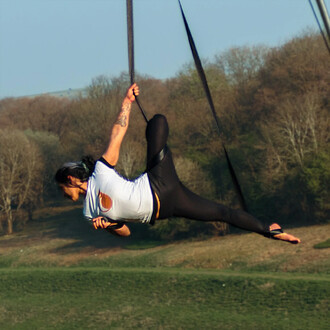 Un artiste acrobatique suspendu dans les sangles aériennes, effectuant une figure élégante en appui sur une main, avec le corps en extension. L'arrière-plan montre un paysage naturel avec des arbres et une prairie verte, sous un ciel clair. L'acrobate est