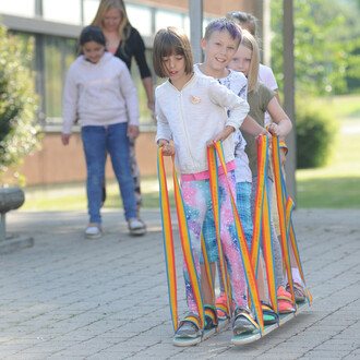 Quatre enfants s'amusent à marcher ensemble sur des grandes échasses en bois. Ils tiennent des cordes multicolores et semblent concentrés tout en avançant. Une femme se tient à l'arrière, observant les enfants, tandis qu'une autre fille est légèrement en 