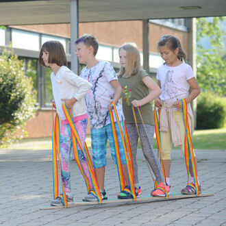 Quatre enfants se tiennent sur une plateforme en bois à l'extérieur, utilisant des strapontins colorés pour avancer ensemble. Ils portent des vêtements colorés et semblent s'amuser tout en essayant de coordonner leurs mouvements.