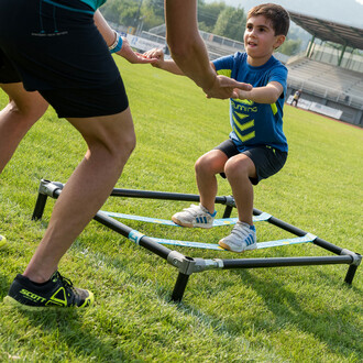 Enfant effectuant un exercice de squat sur une structure autoportante SlackGYM en extérieur, aidé par un adulte, démontrant l'utilisation de l'équipement pour améliorer l'équilibre et la coordination dès le plus jeune âge.