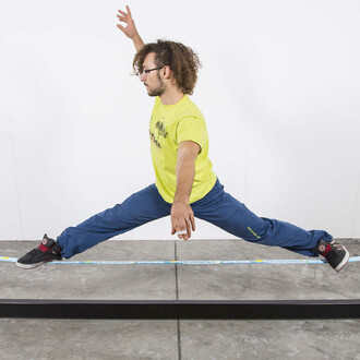 Image montrant une personne effectuant un grand écart sur une slackline, soutenue par la structure Spider Slacklines House 3.0. La personne porte un t-shirt jaune avec le logo Spider Slacklines et un pantalon bleu, démontrant flexibilité et contrôle. La s