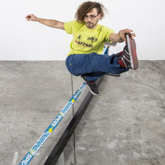 Image montrant une personne effectuant une figure acrobatique sur une slackline, soutenue par la structure Spider Slacklines House 3.0. La personne porte un t-shirt jaune avec le logo Spider Slacklines et un pantalon bleu, les jambes pliées et les bras te