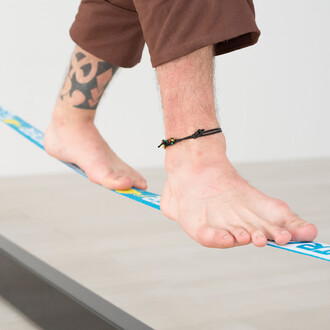 Image montrant les pieds d'une personne en équilibre sur une slackline, soutenue par la structure Spider Slacklines House 3.0. La personne est pieds nus, avec une cheville ornée d'un bracelet noir. La slackline est installée dans un environnement intérieu