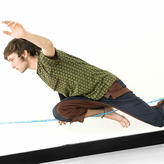 Image montrant une personne effectuant une figure acrobatique sur une slackline, soutenue par la structure Spider Slacklines House 3.0. La personne est en position accroupie, les bras tendus pour maintenir l'équilibre. Elle porte un t-shirt vert à motifs 