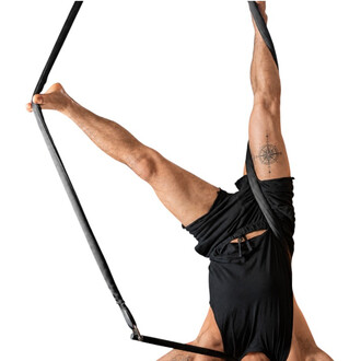 Homem sem camisa, vestindo roupas pretas, suspenso de cabeça para baixo por uma fita Smart Rope preta, com a perna estendida segurando a fita com o pé, fundo branco.