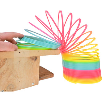 Main tenant un arc-en-ciel géant en plastique coloré, s'étendant depuis un support en bois. L'arc est composé de plusieurs spirales de différentes couleurs, allant du vert au rose, en passant par le jaune et l'orange, créant un effet visuel attrayant.