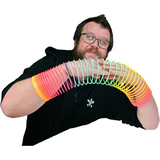 Un homme souriant porte un springy arc-en-ciel de taille moyenne, qui s'étend sur ses bras. Le springy, de couleurs vives allant du rose au vert, est positionné comme un accessoire amusant, tandis qu'il pose ses mains dessus. Il est vêtu d'un sweat noir.