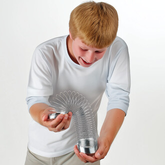 Un enfant souriant tient un ressort en métal brillant, en train de le faire onduler dans les airs. Il porte un t-shirt blanc et des pantalons clairs. L'arrière-plan est neutre, mettant en avant l'objet.