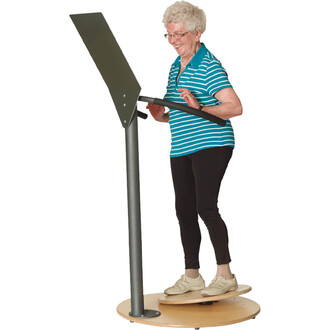 Femme âgée en train d'utiliser un équipement de fitness debout, avec un plateau circulaire en bois et un écran en hauteur, souriante et portant un tee-shirt rayé bleu et blanc. 