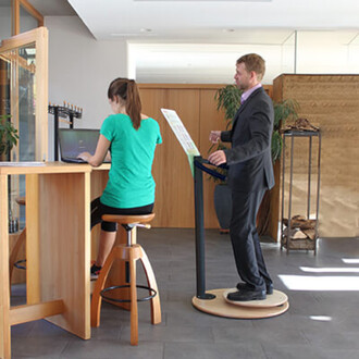 Une image montrant un espace de travail moderne. À gauche, une femme assise sur un tabouret devant un ordinateur portable, vêtue d'un t-shirt vert. À droite, un homme en costume se tient debout sur une plateforme, tenant un document, avec un air attentif.