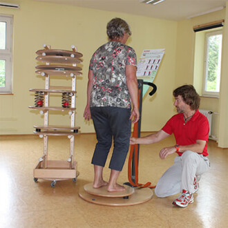 Un homme aide une femme à utiliser un équipement de jonglerie, sur une plateforme rotative. La femme est pieds nus et se tient debout sur la plateforme, tandis que l'homme, à genoux, ajuste un strap. En arrière-plan, un support avec des ressorts et des di