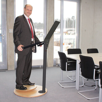 Homme en costume debout sur une plateforme ronde, utilisant un support de présentation. La scène se déroule dans une pièce lumineuse avec de grandes fenêtres. En arrière-plan, une table de réunion avec des chaises noires.