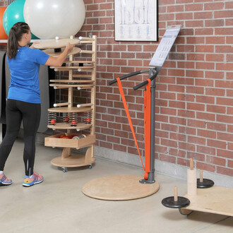 Une personne en tenue de sport utilise un équipement de fitness dans une salle d'exercice. À gauche, un rangement en bois avec des accessoires de fitness, notamment des rouleaux et des poids. À droite, une machine de fitness avec des poignées et une bande