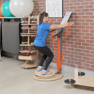 Femme en tenue de sport bleue effectuant un exercice de squat sur un matériel de fitness. Elle utilise des élastiques de résistance fixés à un support mural. En arrière-plan, on distingue des ballons d'exercice, des tapis de sol, et un meuble en bois avec