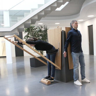 Femme adoptant une posture avant sur un appareil de jonglerie, tenant des barres en bois, tandis qu'un homme se tient en position neutre à côté. L'environnement est moderne avec des escaliers en arrière-plan et une plante verte.