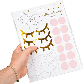 Planche d'autocollants décoratifs yeux blush : cils dorés en plusieurs tailles, cercles roses pastel et motifs lune et étoiles sur fond blanc, tenue par une main.
