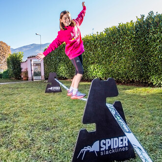 Une jeune fille en sweat-shirt rose pratique la slackline en utilisant des chevalets à trois hauteurs Spider Slacklines dans un jardin ensoleillé, avec des montagnes en arrière-plan