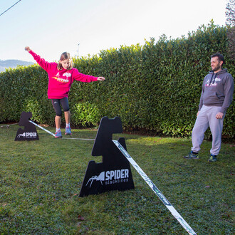 Une jeune fille sur une slackline maintenue par des chevalets à trois hauteurs Spider Slacklines, supervisée par un homme en tenue de sport, dans un jardin spacieux