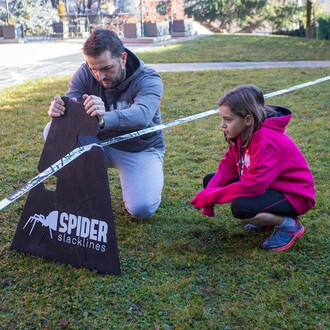 Un homme ajuste un chevalet à trois hauteurs Spider Slacklines tandis qu'une jeune fille l'observe attentivement dans un jardin.