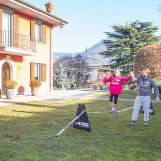 Une famille dans un jardin utilise des chevalets à trois hauteurs Spider Slacklines pour pratiquer la slackline, avec une grande maison en arrière-plan.