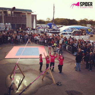  Un événement de tricklining avec un public nombreux. Une slackline est installée sur un support en A en bois, entourée par des tapis de protection au sol. Plusieurs personnes en t-shirts roses de Spider Slacklines posent devant la slackline. En arrière-p