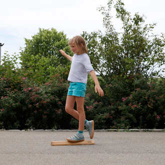 Une jeune fille s'entraîne à marcher sur une barre d'équilibre. Elle porte un t-shirt à rayures blanches et une short turquoise, et se tient en équilibre sur la barre en bois. En arrière-plan, on peut voir des plantes et des arbres, avec un ciel légèremen