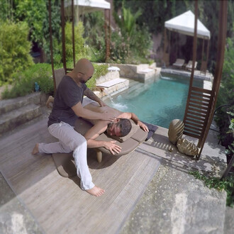 Un homme reçoit un massage relaxant en étant allongé sur une table de massage à l'extérieur, près d'une piscine. L'élément de la piscine est visible en arrière-plan avec des chaises longues. Le masseur, portant des lunettes de soleil et un t-shirt, appliq