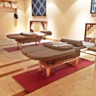 Deux tables de massage en bois, avec des matelas marron et des serviettes blanches, placées sur des tapis rouges dans une salle de massage lumineuse avec des murs clairs et des décorations minimalistes.