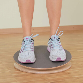 Pieds d'une personne portant des chaussures de sport blanches et bleues sur un disque d'équilibre en bois, sur un sol en bois clair.