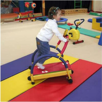 Un enfant s'amuse sur un tapis de marche coloré, utilisant un matériel de jonglerie. Il se tient debout sur un équipement de jeu avec un guidon, sur un tapis au sol composé de différentes couleurs. L'environnement est une salle de sport pour enfants, doté