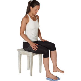 Femme assise sur une chaise en bois, portant un débardeur blanc et un legging noir, utilisant un accessoire de massage sous ses pieds. Elle a une expression détendue et semble profiter de son moment de détente.