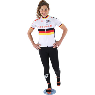 Femme souriante portant un maillot de cyclisme blanc avec des bandes colorées représentant l'Allemagne, et un pantalon noir. Elle se tient sur une plateforme avec une main sur la hanche et l'autre bras légèrement levé.