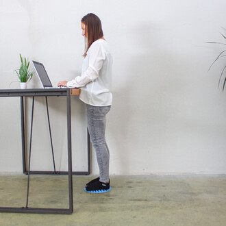 Femme debout à un bureau élevé, utilisant un ordinateur portable, vêtue d'une blouse blanche et de jeans, avec des chaussures noires à semelles bleues. Une plante en pot est posée sur le bureau et le fond est sobre avec des murs clairs.