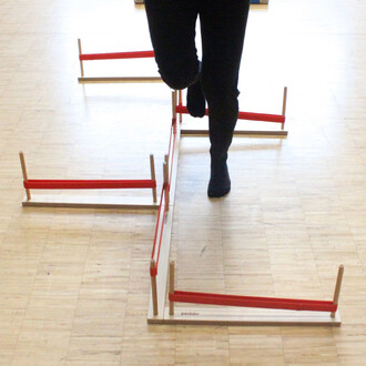 Un individu effectue un exercice en passant le pied au-dessus de barres en bois avec des bandes élastiques rouges, disposées au sol pour un entraînement de coordination, sur un sol en bois.