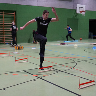 Un athlète s'entraîne dans une salle de sport, sautant par-dessus des haies en bois avec des barres rouges. En arrière-plan, d'autres personnes s'exercent avec divers équipements de fitness. Le sol est en synthétique, et des lignes de marquage sont visibl