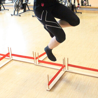 Une personne en train de sauter au-dessus d'un obstacle de saut en hauteur, constitué de barres en bois et de lanières rouges, sur un sol en bois dans un environnement de salle de sport.