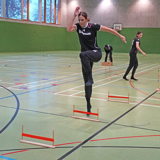 Une athlète s'apprête à sauter par-dessus des haies basses dans une salle de sport. Elle porte un t-shirt noir et un pantalon noir. Deux autres personnes sont visibles en arrière-plan, l'une en train de placer des obstacles et l'autre se déplaçant sur le 