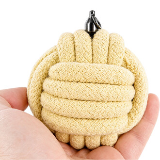 Monkey fist en corde beige, nœud sphérique serré avec attache métallique et anneau au sommet, tenu entre les doigts.