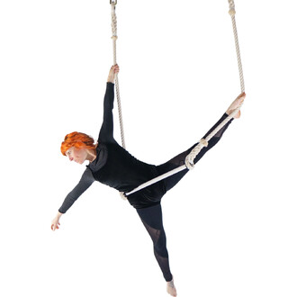 Artiste de cirque exécutant une figure acrobatique sur un trapèze classique en coton, suspendue par des cordes. Elle porte un vêtement noir et a les cheveux roux, avec une jambe étendue et les bras en position équilibrée.