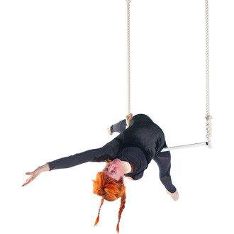 Femme en position acrobatique sur un trapèze classique en coton, suspendue par des cordes blanches, avec des cheveux roux en tresses et portant un costume noir ajusté.