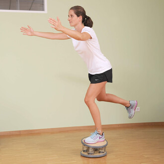 Femme en tenue de sport, réalisant un exercice d'équilibre sur une plateforme de jonglage, avec les bras tendus et une jambe levée, sur un sol en parquet clair.