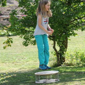 Enfant sautant sur un tremplin Pedalo® 500, vêtu d'un débardeur gris et de pantalons turquoise, dans un jardin avec un arbre en arrière-plan.