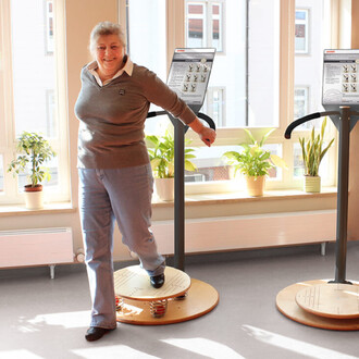 Personne souriante utilisant un équipement de fitness sur une plateforme oscillante, à l'intérieur d'une pièce lumineuse avec des plantes. Des panneaux d'instructions sont visibles de chaque côté.