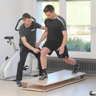 Un entraîneur aide un jeune homme à réaliser un exercice d'équilibre sur une plateforme, dans une salle d'entraînement bien éclairée. L'entraîneur, installé à côté de l'appareil de fitness, guide le jeune homme qui est en position d'un pied sur la platefo