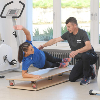 Femme en position de planche latérale sur un banc de fitness, avec un entraîneur qui l'aide en lui tenant les hanches. Le décor est une salle de sport lumineuse, avec du matériel de fitness en arrière-plan.