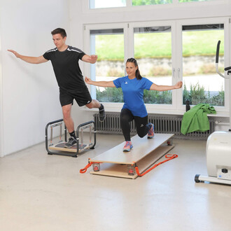 Un homme en tenue de sport noire effectue un exercice d'équilibre sur une plateforme, tandis qu'une femme en t-shirt bleu fait un exercice dynamique sur une planche de gymnastique. L'environnement est lumineux avec des fenêtres donnant sur l'extérieur et 